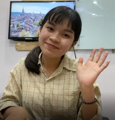 A young woman, identified as a KL escort girl, is sitting at a desk with her hand raised.