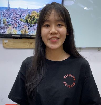 An Asian woman in a black t-shirt standing in front of a TV.